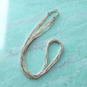 Op de afbeelding: Een zilveren ketting met meerdere strengen, bestaande uit talloze dunne, delicate metalen kettinkjes die aan de bovenkant zijn samengevoegd met een kleine zilveren sluiting. De ketting is in een lange, vloeiende lus gelegd, wat het reflecterende, gepolijste oppervlak van het metaal benadrukt.