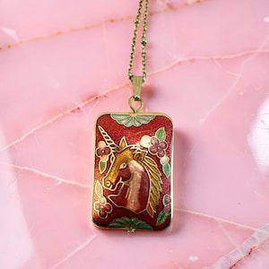 Puede incluir: Collar con colgante rectangular. El colgante tiene un fondo rojo con un diseño de unicornio dorado y detalles florales. El colgante está enmarcado con un borde dorado.
