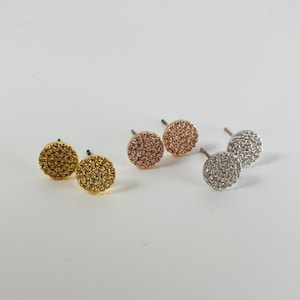May include: Three pairs of small, round stud earrings. The earrings are made of metal and have a sparkly, textured surface. One pair is gold, one is rose gold, and one is silver.