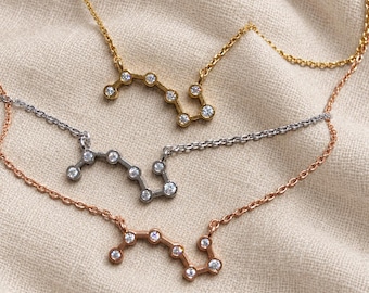 May include: Three delicate necklaces featuring a constellation pendant design. Each pendant is set with small, clear round stones arranged in a zig-zag pattern. The necklaces are available in gold, silver, and rose gold metal finishes with fine chains.