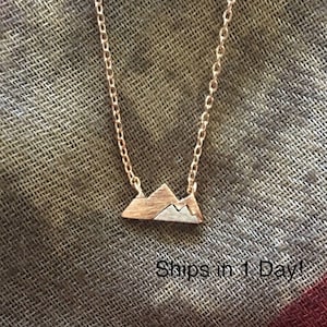May include: A delicate gold chain necklace with a small, two-toned mountain pendant. The mountain is made of rose gold and silver, with a textured finish.