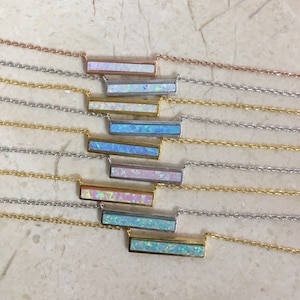 May include: A set of nine necklaces with bar pendants featuring iridescent opal stones in various shades of blue, white, and pink. The necklaces are layered on a white textured surface.
