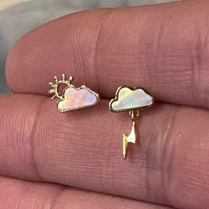 May include: A pair of gold-colored earrings. One earring features a cloud with a sun, and the other has a cloud with a dangling lightning bolt. The clouds have an iridescent, opalescent appearance.