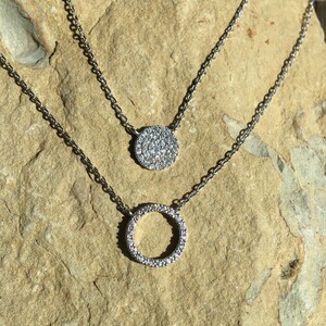 May include: Two silver necklaces with pendant designs. The top necklace features a circular pendant covered in small, clear stones. The bottom necklace has a circle pendant also covered in clear stones. The necklaces are displayed on a light brown stone surface.