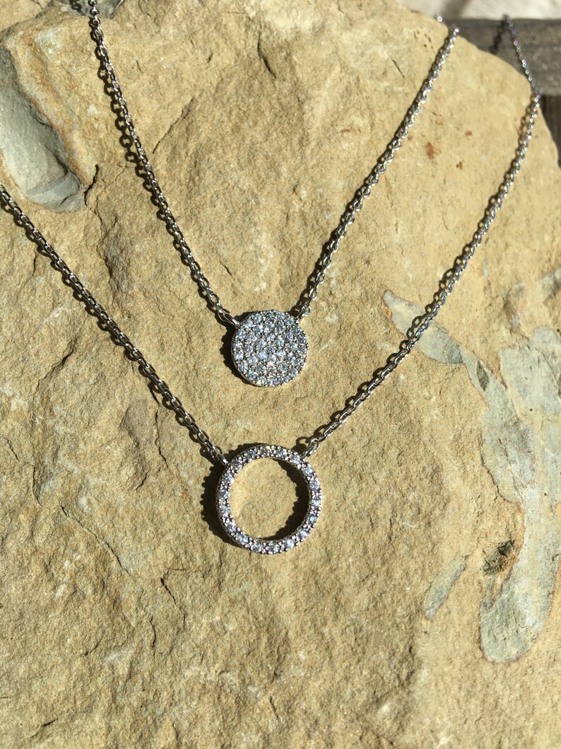 May include: Two silver necklaces with pendant designs. The top necklace features a circular pendant covered in small, clear stones. The bottom necklace has a circle pendant also covered in clear stones. The necklaces are displayed on a light brown stone surface.