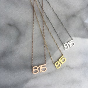 May include: Three gold necklaces with the number 815 hanging from them. The necklaces are on a white marble surface. The necklaces are in different shades of gold, including rose gold, yellow gold, and silver.