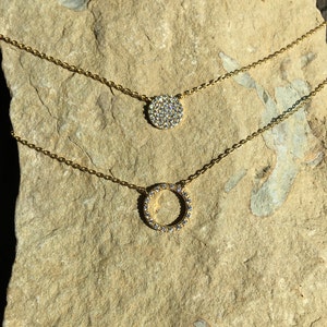 May include: Two gold-colored necklaces with pendant designs. The top necklace features a round pendant covered in small, clear stones. The bottom necklace has a circular pendant also adorned with clear stones. Both necklaces rest on a light brown, textured surface.