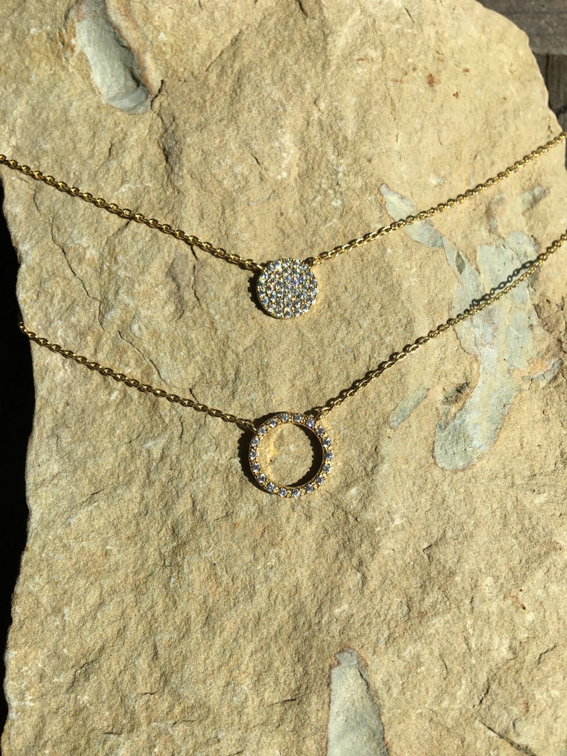 May include: Two gold-colored necklaces with pendant designs. The top necklace features a round pendant covered in small, clear stones. The bottom necklace has a circular pendant also adorned with clear stones. Both necklaces rest on a light brown, textured surface.