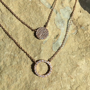 May include: Two rose gold necklaces with delicate chains. The top necklace features a small, round pendant fully encrusted with sparkling crystals. The bottom necklace has a larger open circle pendant, also adorned with crystals.