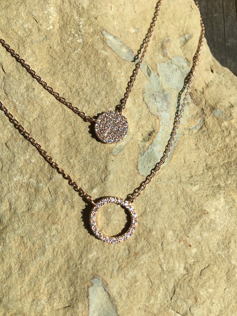 May include: Two rose gold necklaces with delicate chains. The top necklace features a small, round pendant fully encrusted with sparkling crystals. The bottom necklace has a larger open circle pendant, also adorned with crystals.