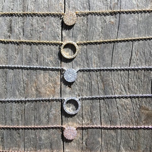 May include: A collection of delicate necklaces with circular pendants. The pendants are encrusted with small, sparkling stones. The chains and pendants come in gold, silver, and rose gold tones, displayed on a weathered wooden surface.