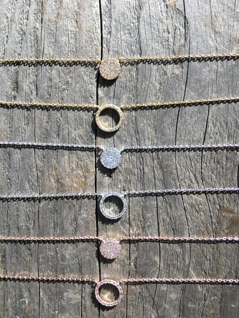 May include: A collection of delicate necklaces with circular pendants. The pendants are encrusted with small, sparkling stones. The chains and pendants come in gold, silver, and rose gold tones, displayed on a weathered wooden surface.