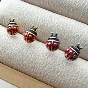 May include: Four stud earrings shaped like ladybugs, each featuring a red body with black spots and a black head. The earrings are crafted with a glossy finish and metallic accents, displaying a rounded, insect-inspired design for ear adornment.