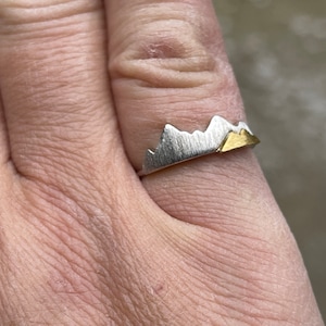May include: A silver ring with a gold mountain peak design. The ring is worn on a finger.