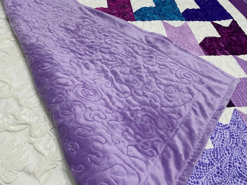May include: A soft, purple blanket with a swirling pattern. The blanket is folded over a white and purple quilt.