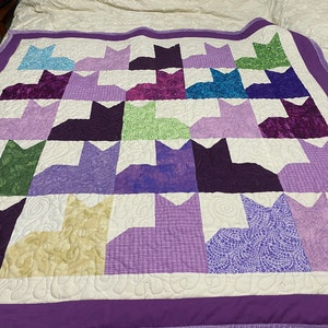 May include: A purple and white quilt with a pattern of 24 purple, green, and cream cats. The quilt is quilted with a swirl pattern.