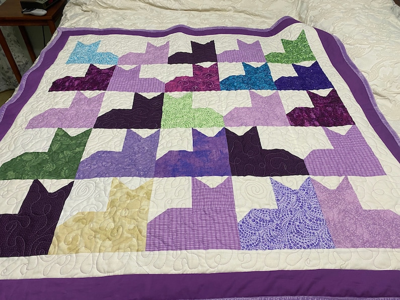 May include: A purple and white quilt with a pattern of 24 purple, green, and cream cats. The quilt is quilted with a swirl pattern.