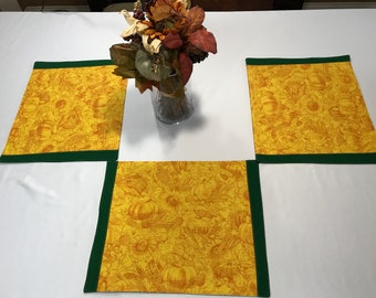 May include: Four rectangular placemats with a yellow and orange autumn-themed pattern featuring pumpkins and leaves. Each placemat has a dark green border. A vase of fall foliage sits in the center.