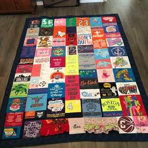 Op de afbeelding: Een patchworkdeken gemaakt van verschillende T-shirts. De deken is blauw met een verscheidenheid aan kleuren en ontwerpen. De deken bevat tekst zoals "Hester 23", "Conquerors Don't Quit", "Play", "Be Kind", "Faith Works!" en "New York City".