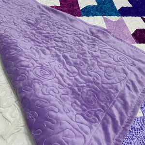 May include: A soft, purple blanket with a swirling pattern. The blanket is folded over a white and purple quilt.