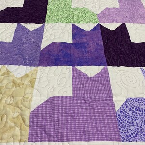 May include: A quilt with a patchwork design featuring various shades of purple, green, and beige. The quilt is made up of squares with different patterns, including polka dots, plaid, and floral designs.