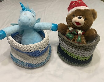 May include: Two handmade crochet baskets, one holding a blue unicorn plush toy and the other a brown teddy bear wearing a Santa hat and scarf. The baskets are in shades of blue, gray, and green.