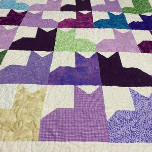 May include: A purple, green, and white quilt with a pattern of cats. The quilt is made of different fabrics with various textures and patterns.