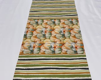May include: A long, rectangular table runner with a striped pattern in shades of green, brown, and black. The center of the runner features a pattern of white and orange pumpkins.