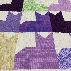 May include: A quilt with a patchwork design featuring purple, green, and beige fabric. The quilt is made up of squares with different patterns and colors. The quilt is quilted with a white thread.
