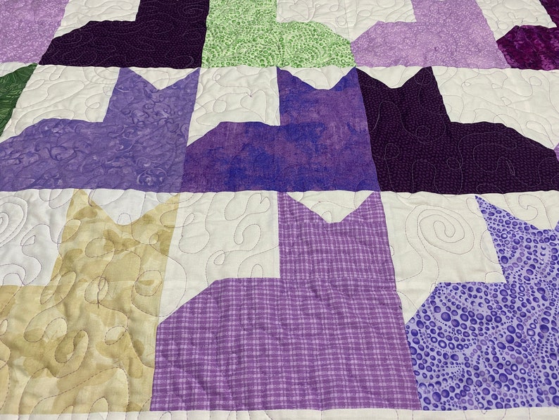 May include: A quilt with a patchwork design featuring purple, green, and beige fabric. The quilt is made up of squares with different patterns and colors. The quilt is quilted with a white thread.