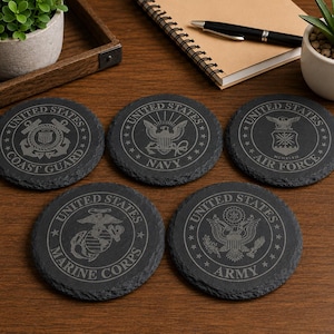 May include: Five dark grey slate coasters, each with a different etched emblem of a United States military branch. The coasters display the Coast Guard, Navy, Air Force, Marine Corps, and Army insignias, ideal for drinks.