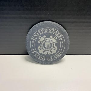 May include: A circular, grey slate coaster featuring the text "UNITED STATES COAST GUARD" around a central emblem. The emblem includes an anchor, rope, and shield design, all in a lighter grey. The coaster rests on a white surface.