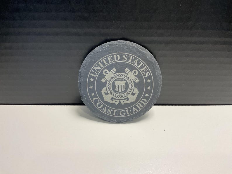 May include: A circular, grey slate coaster featuring the text "UNITED STATES COAST GUARD" around a central emblem. The emblem includes an anchor, rope, and shield design, all in a lighter grey. The coaster rests on a white surface.