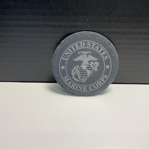 May include: A circular, grey slate coaster featuring the words "United States Marine Corps" engraved around the edge. The centre displays the Marine Corps emblem. The coaster is on a white surface.