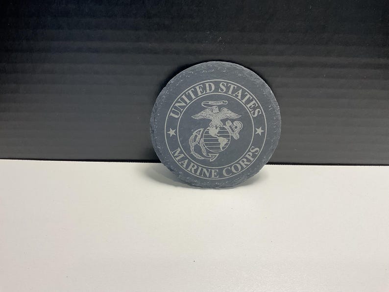 May include: A circular, grey slate coaster featuring the words "United States Marine Corps" engraved around the edge. The centre displays the Marine Corps emblem. The coaster is on a white surface.