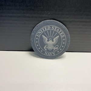 May include: A circular, grey slate coaster featuring the words "UNITED STATES NAVY" etched around a central emblem of an eagle and shield. The coaster is a dark grey colour and has a natural, textured surface.