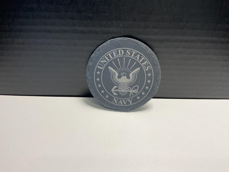 May include: A circular, grey slate coaster featuring the words "UNITED STATES NAVY" etched around a central emblem of an eagle and shield. The coaster is a dark grey colour and has a natural, textured surface.