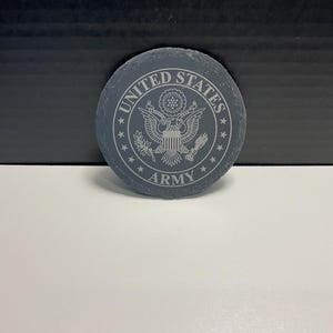 May include: A circular, dark grey stone coaster featuring the words "UNITED STATES ARMY" around a detailed emblem. The emblem includes an eagle, stars, and other intricate designs. The coaster is on a white surface.