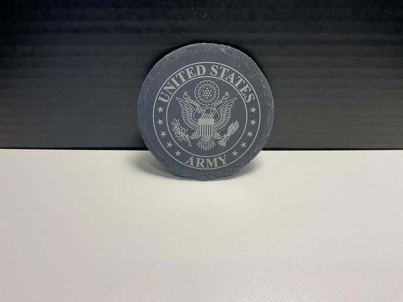 May include: A circular, dark grey stone coaster featuring the words "UNITED STATES ARMY" around a detailed emblem. The emblem includes an eagle, stars, and other intricate designs. The coaster is on a white surface.