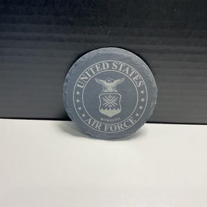 May include: A circular, grey slate coaster featuring the United States Air Force emblem. The design includes an eagle, shield, and the words "United States Air Force" and "MCMXLVII" around the edge. The coaster is approximately 10 cm in diameter.