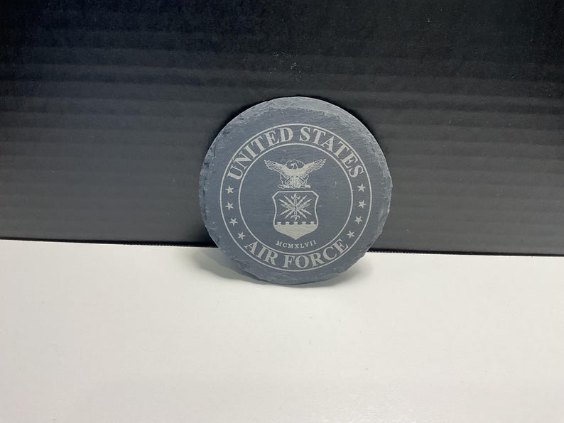 May include: A circular, grey slate coaster featuring the United States Air Force emblem. The design includes an eagle, shield, and the words "United States Air Force" and "MCMXLVII" around the edge. The coaster is approximately 10 cm in diameter.