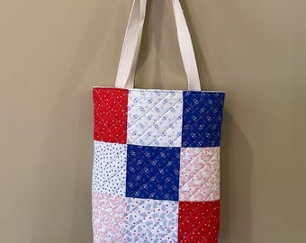 May include: Quilted tote bag with a patchwork design in red, white, and blue. The bag features a variety of patterns, including polka dots, floral prints, and geometric shapes. It has cream-colored straps and a decorative scalloped edge.