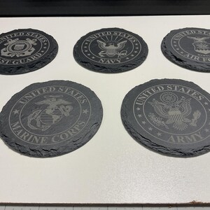 May include: Five circular, dark grey slate coasters with etched designs. Each coaster features a different emblem representing branches of the United States military: Coast Guard, Navy, Air Force, Marine Corps, and Army.