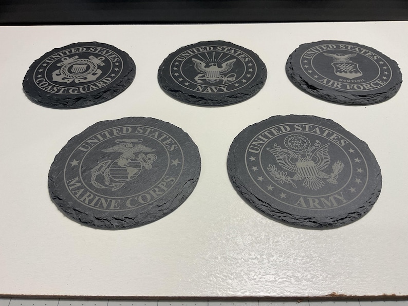 May include: Five circular, dark grey slate coasters with etched designs. Each coaster features a different emblem representing branches of the United States military: Coast Guard, Navy, Air Force, Marine Corps, and Army.