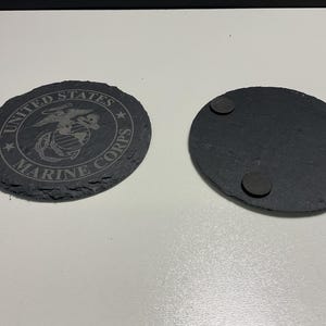 May include: Two black slate coasters. One coaster features the United States Marine Corps emblem and text. The other coaster has three small, round, dark grey pads on the back. The coasters are oval-shaped with a rough, natural edge.