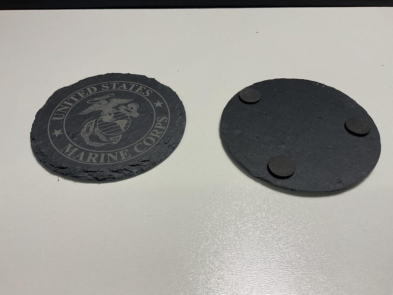 May include: Two black slate coasters. One coaster features the United States Marine Corps emblem and text. The other coaster has three small, round, dark grey pads on the back. The coasters are oval-shaped with a rough, natural edge.