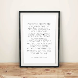 May include: A framed print with a black border, featuring a quote by Sir Arthur Conan Doyle in black text on a white background. The quote encourages taking a bicycle ride. The frame is leaning against a light-colored wall and a wooden floor.