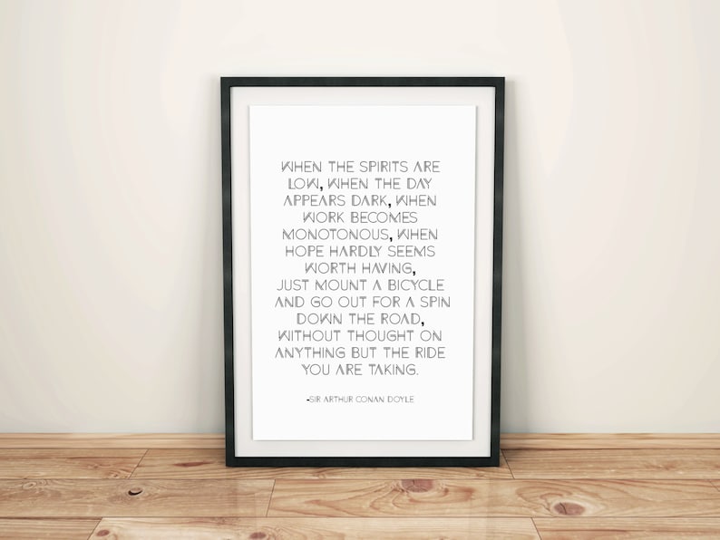 May include: A framed print with a black border, featuring a quote by Sir Arthur Conan Doyle in black text on a white background. The quote encourages taking a bicycle ride. The frame is leaning against a light-colored wall and a wooden floor.
