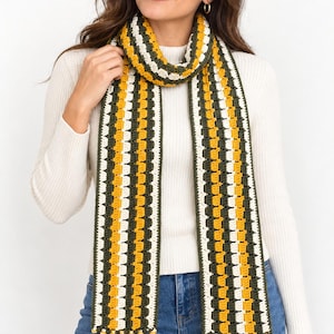 May include: A long crochet scarf featuring vertical stripes in mustard yellow, forest green, and cream white. The scarf is constructed with a textured stitch pattern and finished with long, multi-coloured fringe tassels at both ends of the fabric.