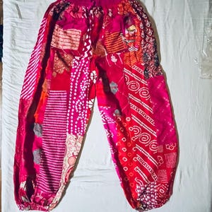 Könnte beinhalten: Helle, rosafarbene Patchworkhose mit Kordelzug. Die Hose zeigt verschiedene Muster, darunter Streifen, Punkte und geometrische Designs in Rot, Rosa und Weiß. Die Hose hat eine entspannte Passform im Harem-Stil.